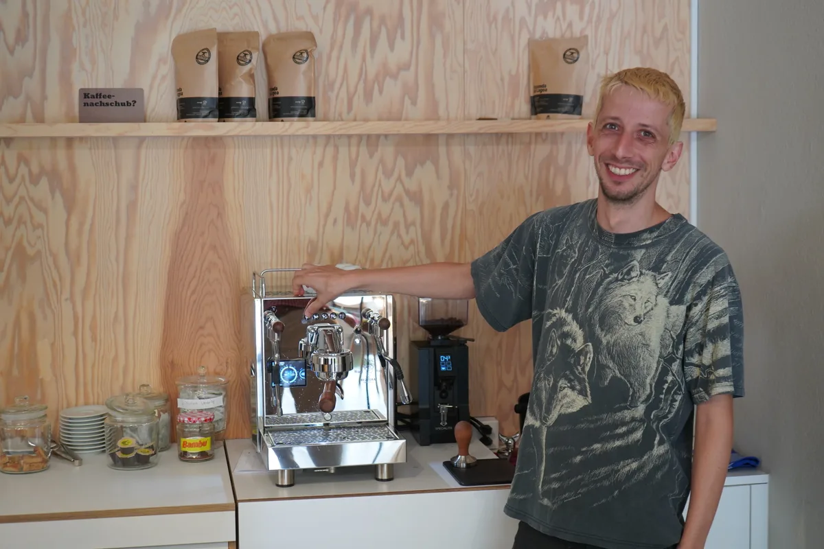 Ein junger Mann strahlt neben einer Kaffeemaschine in die Kamera.
